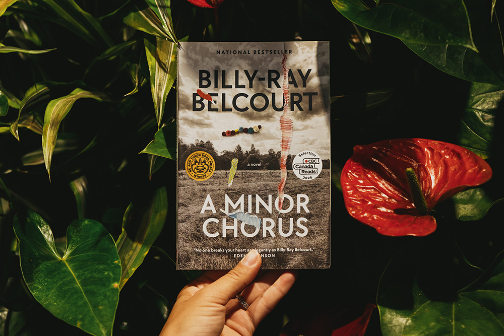 A hand holds a book titled "A Minor Chorus, By Billy-Ray Belcourt" against a backdrop of lush plants